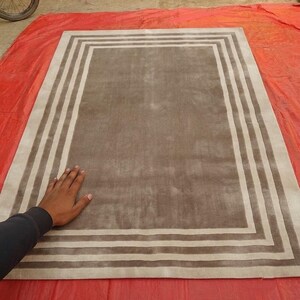 May include: A rectangular area rug featuring a solid taupe center panel surrounded by a series of concentric rectangular borders in alternating shades of cream and taupe, creating a clean, geometric frame design across the entire surface.
