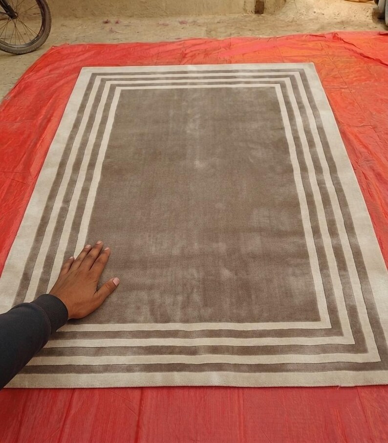 May include: A rectangular area rug featuring a solid taupe center panel surrounded by a series of concentric rectangular borders in alternating shades of cream and taupe, creating a clean, geometric frame design across the entire surface.