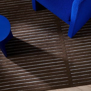May include: A dark brown area rug featuring a pattern of thin, horizontal white stripes. The rug is positioned beneath a vibrant blue armchair and a matching blue circular side table, creating a high-contrast visual arrangement of furniture.