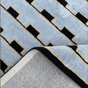 K&ouml;nnte beinhalten: Ein rechteckiger Teppich mit einem sich wiederholenden geometrischen Muster aus hellblauen Bl&ouml;cken, die durch schwarze rechteckige Formen und d&uuml;nne beige Umrandungen getrennt sind. Eine Ecke ist umgeschlagen und zeigt eine hellgraue R&uuml;ckseite.