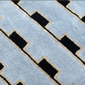 K&ouml;nnte beinhalten: Ein strukturierter Teppich mit einem sich wiederholenden geometrischen Muster aus hellblauen rechteckigen Formen, die in einer versetzten, ziegelartigen Anordnung angeordnet sind. Jeder hellblaue Bereich ist mit einem cremefarbenen Rand abgesetzt.