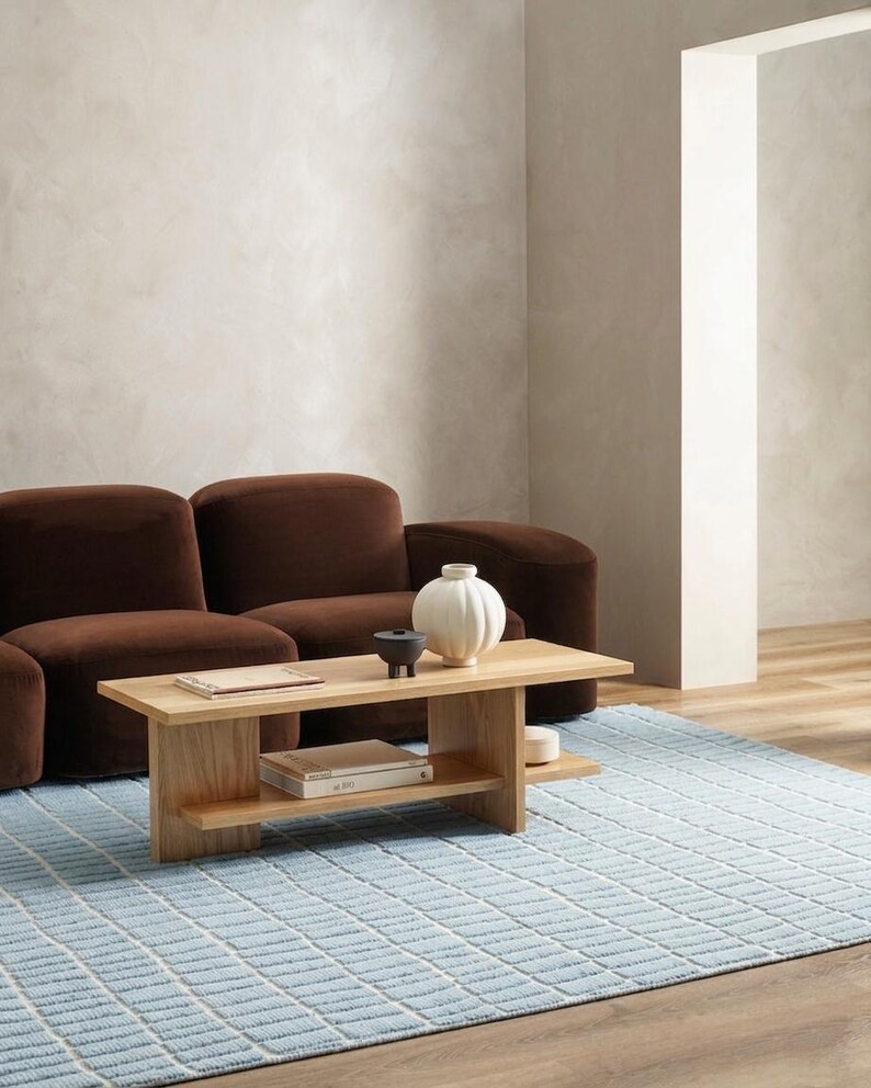 May include: A modern living room scene featuring a brown sofa, a light wood coffee table with a lower shelf, and a light blue patterned rug. Decorative items on the table include a white vase and a black bowl. The room has a neutral color palette.