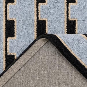 K&ouml;nnte beinhalten: Ein getufteter Teppich mit hellblauem Hintergrund und einem geometrischen Muster aus schwarzen vertikalen Linien und d&uuml;nnen beigen R&auml;ndern. Die Ecke des Teppichs ist umgeschlagen und zeigt eine schlichte graue Stoffr&uuml;ckseite sowie eine vern&auml;hte Kante.