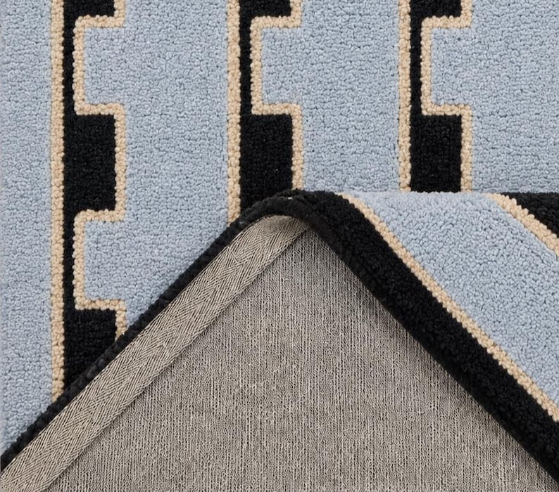 K&ouml;nnte beinhalten: Ein getufteter Teppich mit hellblauem Hintergrund und einem geometrischen Muster aus schwarzen vertikalen Linien und d&uuml;nnen beigen R&auml;ndern. Die Ecke des Teppichs ist umgeschlagen und zeigt eine schlichte graue Stoffr&uuml;ckseite sowie eine vern&auml;hte Kante.