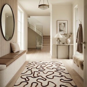 Può includere: Un tappeto rettangolare con base color crema e un motivo a linee astratte continue e sinuose di colore marrone scuro. Il tappeto è posizionato in un corridoio con una panca integrata, una console, uno specchio e una lampada a sospensione.