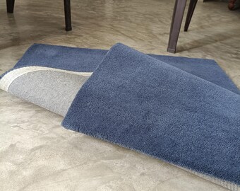 Puede incluir: Una alfombra rectangular con una superficie de pelo azul profundo y un reverso tejido en gris claro. La alfombra está doblada por una esquina, revelando el contraste entre la suave textura azul superior y el material estructurado de color claro.