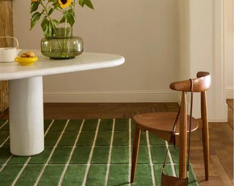 May include: A green rectangular rug with white lines, a white round table, and a wooden chair. A vase of sunflowers sits on the table. A brown leather bag hangs on the chair.