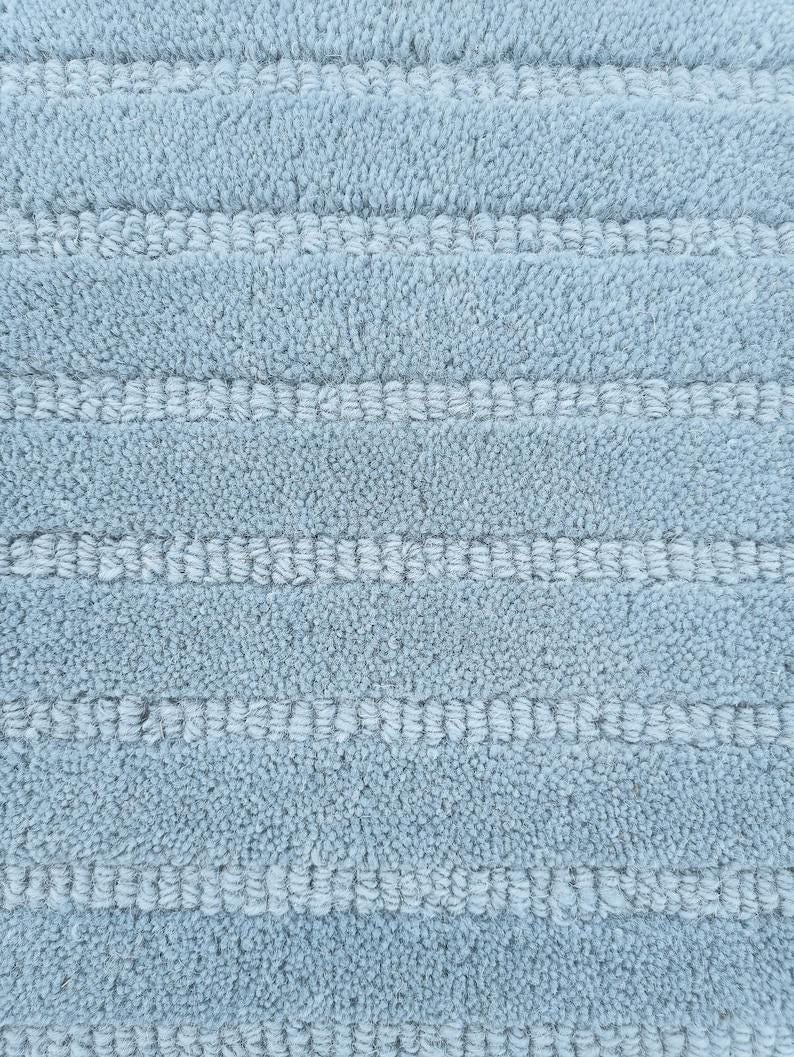 May include: Close-up of a light blue bath mat with a textured, looped pile. The mat features horizontal stripes of a lighter blue color, creating a subtle pattern. The soft, plush material suggests comfort and absorbency.