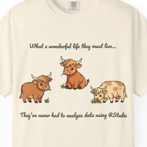 May include: Cream-colored t-shirt featuring a whimsical illustration of three cartoon Highland cows. The cows are in shades of brown and tan, with small tufts of grass beneath them. Text reads, "What a wonderful life they must live..." and "They've never had to analyze data using RStudio."