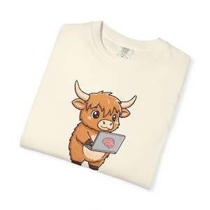 May include: Cream-colored t-shirt featuring a cartoon Highland cow holding a laptop with a brain graphic on the screen. The cow is brown with large horns and a fluffy coat. The shirt is folded.