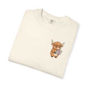May include: Cream-colored t-shirt featuring a cartoon Highland cow holding a laptop with a brain graphic on the screen. The shirt is folded, with the Comfort Colors label visible in the upper corner.