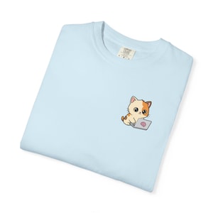 Pode incluir: Camiseta azul clara com um gato de desenho animado com pelo laranja e branco e um laptop. O gato está sentado e parece estar trabalhando no laptop. A camiseta tem gola redonda e uma etiqueta por dentro.