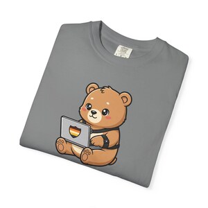 Può includere: T-shirt grigio con un orsetto dei cartoni animati che usa un laptop. L'orso è marrone con un'imbracatura nera e tiene in mano un laptop con un design arcobaleno. La maglietta ha un'etichetta Comfort Colors.
