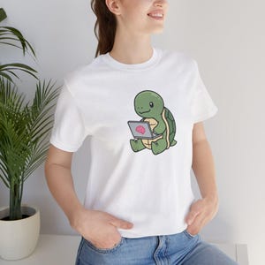 Könnte beinhalten: Weißes T-Shirt mit einer Cartoon-Schildkröte, die einen Laptop benutzt. Die Schildkröte ist grün mit einem beigen Bauch und einem dunkelgrünen Panzer. Der Laptop-Bildschirm zeigt ein rosa Gehirn. Das T-Shirt hat einen Rundhalsausschnitt.