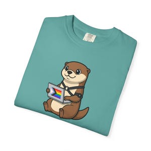 Könnte beinhalten: Türkisfarbenes T-Shirt mit einer Cartoon-Otter, die einen Laptop mit einer Regenbogen- und Transgender-Pride-Flagge hält. Der Otter ist braun mit schwarzen Akzenten. Das Shirt hat einen Rundhalsausschnitt und ein kleines Etikett oben rechts.