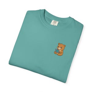 May include: A teal Comfort Colors t-shirt featuring a small embroidered graphic of a brown teddy bear holding a laptop. The bear is sitting and the laptop screen displays a brain. The shirt is folded and has a tag.