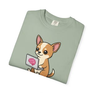 Könnte beinhalten: Salbeigrünes T-Shirt mit einem Cartoon-Chihuahua. Der Hund mit braun-weißem Fell hält einen Laptop mit einer rosa Gehirn-Grafik. Das T-Shirt hat einen Rundhalsausschnitt und ein kleines Etikett oben rechts.