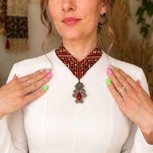 May include: A V-shaped choker necklace featuring intricate red, black, and yellow geometric embroidery. A silver-toned metal pendant hangs from the center, decorated with ornate filigree swirls and a single smooth, teardrop-shaped red stone.
