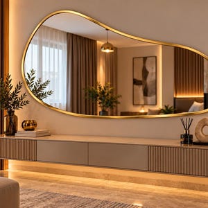 May include: An irregular, organic-shaped wall mirror with a thin gold frame mounted above a long, floating media console. The console features a combination of smooth and vertically slatted panels in a neutral beige tone, illuminated by integrated warm LED lighting.