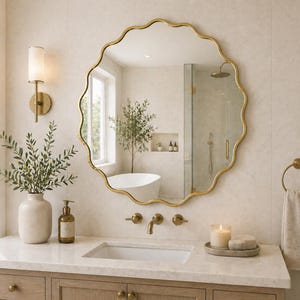 May include: A gold-framed, scalloped-edge mirror hangs above a bathroom vanity. The vanity features a white countertop, a sink, and a wooden cabinet. A vase with greenery, a soap dispenser, and a candle are on the counter.
