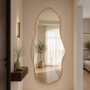 Op de afbeelding: Een onregelmatig gevormde spiegel met een gouden frame hangt aan een muur in neutrale tinten. De spiegel weerspiegelt een kamer met een boom, gordijnen en zonlicht. Een klein plankje met een vaas en groen staat links van de spiegel.