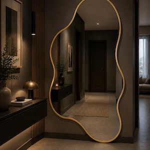 Op de afbeelding: Een grote, goudomrande, vrijvormige spiegel hangt aan een donkergrijze muur in een gang. De spiegel reflecteert de gang, een donkere houten deur en een tapijt in neutrale tinten. De gang heeft een modern, minimalistisch design.