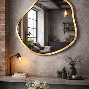 Op de afbeelding: Een grote, onregelmatig gevormde spiegel met een goudkleurige lijst hangt aan een grijze betonnen muur. De spiegel reflecteert een kamer met een bank, een raam en een zwarte bureaulamp. Een tafel met een vaas bloemen staat op de voorgrond.