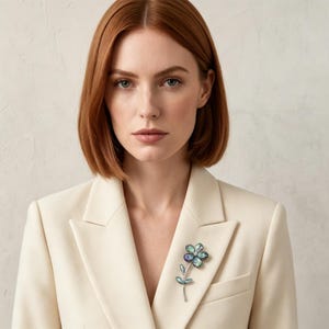 Puede incluir: Un broche con forma de flor con detalles azules, verdes y morados está prendido a una blazer color crema. El broche tiene un tallo y hojas plateadas. La mujer de la imagen tiene el pelo rojo y lleva una blazer color crema.
