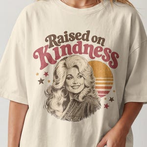 May include: An off-white short-sleeve t-shirt featuring a graphic print of a woman with voluminous hair. The text reads Raised on Kindness in brown and red letters. A circular orange and yellow sunset graphic and small stars surround the portrait.