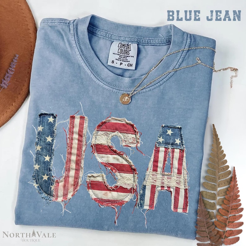 May include: A blue jean colored t-shirt with the word "USA" appliqued on the front in a distressed American flag design. A gold necklace with a pendant rests on the shirt. The shirt is folded and displayed with a hat and dried leaves.
