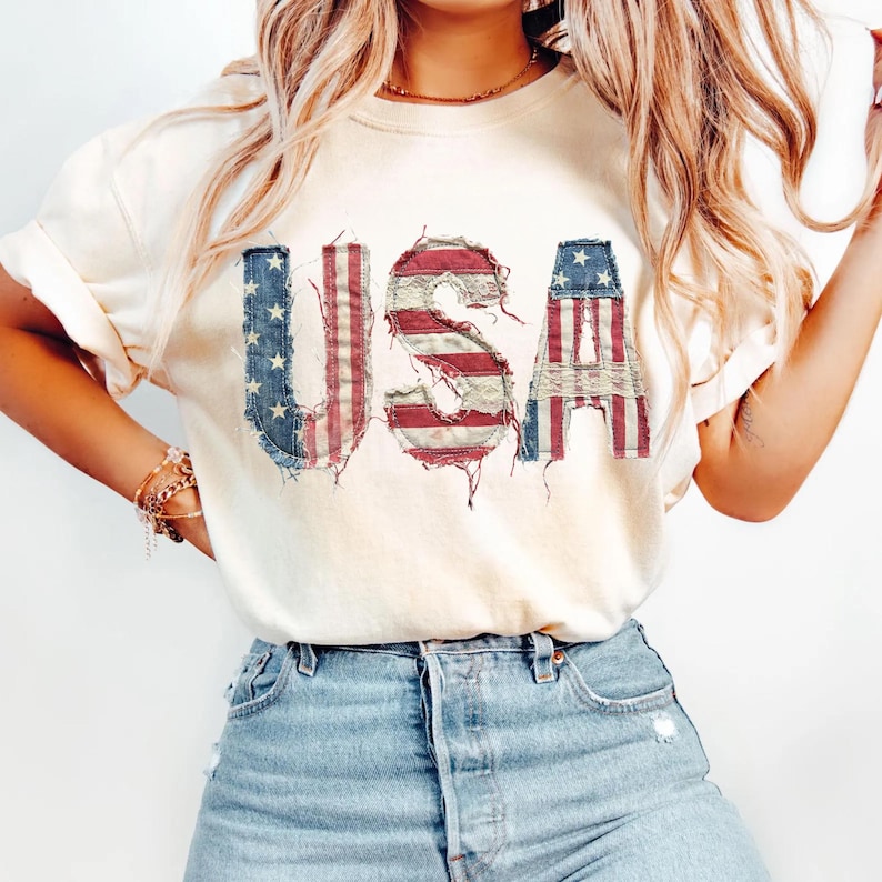 May include: Cream-colored t-shirt with the word "USA" in distressed, patriotic lettering. The letters feature a patchwork design with red and white stripes and blue stars, evoking the American flag. The shirt is paired with light blue denim jeans.