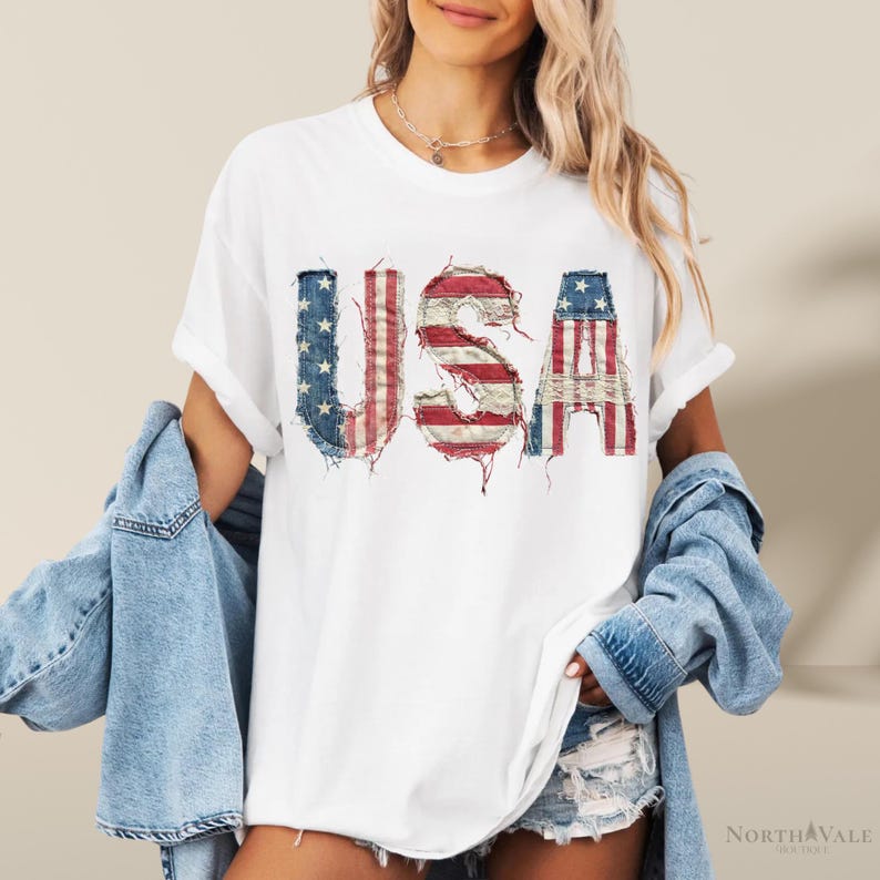 May include: White t-shirt with the word "USA" in distressed red, white, and blue fabric, resembling the American flag. The shirt has rolled-up sleeves and is worn with a denim jacket and shorts.
