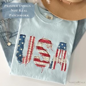 May include: Light blue t-shirt with the word "USA" in a patchwork design featuring the American flag. The shirt is folded on top of a pair of blue jeans. A tan hat and a necklace are also visible.