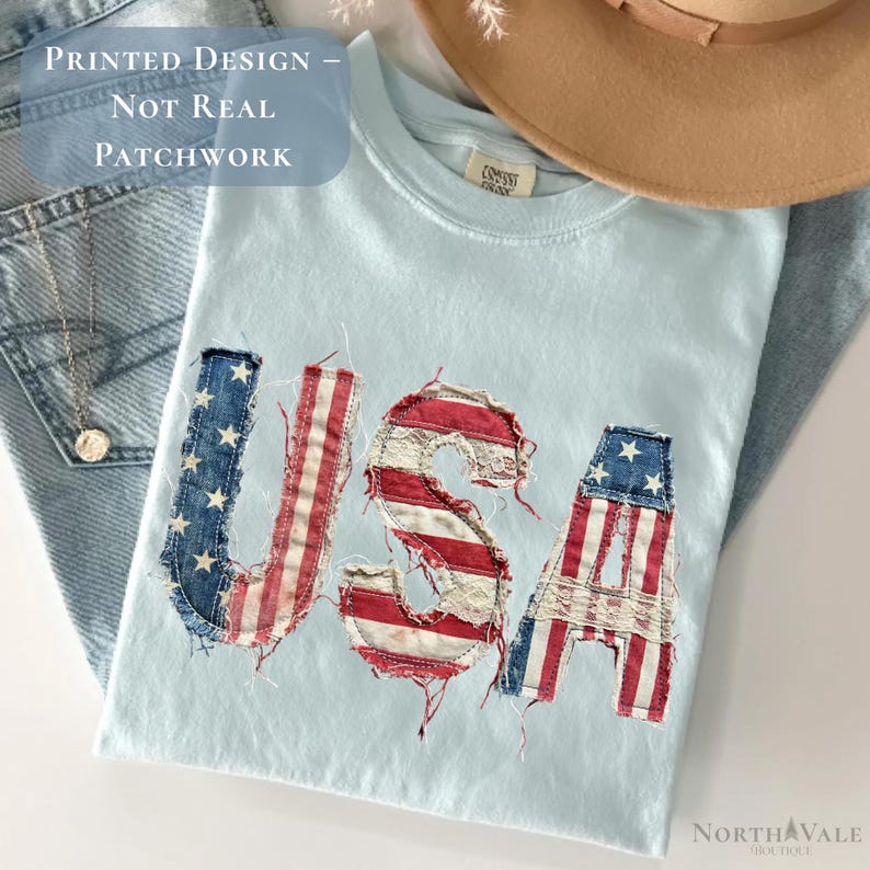 May include: Light blue t-shirt with the word "USA" in a patchwork design featuring the American flag. The shirt is folded on top of a pair of blue jeans. A tan hat and a necklace are also visible.