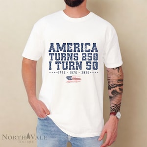 May include: White t-shirt with the text "AMERICA TURNS 250 I TURN 50" in distressed blue lettering. Below the text is a small American flag graphic and the years 1776-1976-2026. The shirt is short-sleeved.