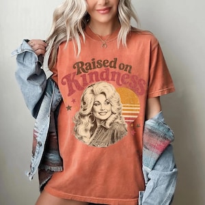 May include: A burnt orange short-sleeve t-shirt featuring a graphic print of a blonde woman with voluminous hair. The text "Raised on Kindness" is arched above the portrait, accompanied by a yellow sun graphic and small star icons.