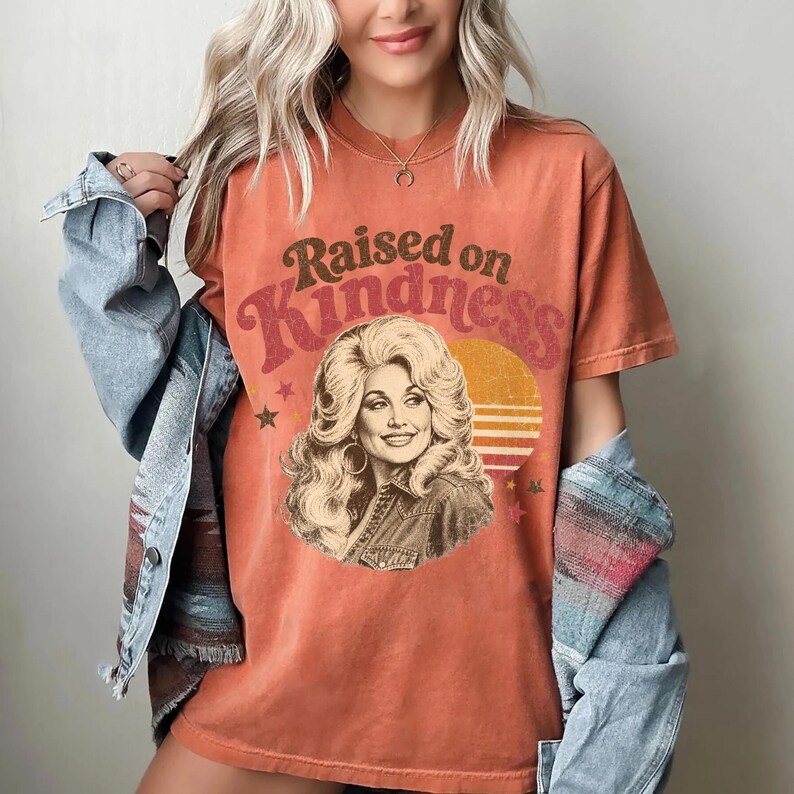May include: A burnt orange short-sleeve t-shirt featuring a graphic print of a blonde woman with voluminous hair. The text "Raised on Kindness" is arched above the portrait, accompanied by a yellow sun graphic and small star icons.