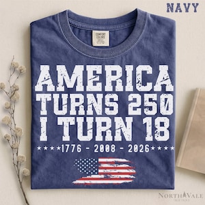 May include: A navy blue crew neck t-shirt featuring distressed white text that reads AMERICA TURNS 250 I TURN 18, followed by the years 1776 - 2008 - 2026 and a graphic of a distressed American flag at the bottom center of the garment.
