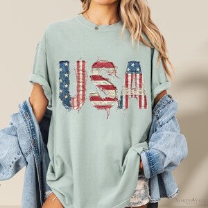 May include: A light green t-shirt with the word "USA" in a distressed, patriotic design. The letters are made to look like the American flag, with red and white stripes and blue stars. The shirt has rolled-up sleeves.