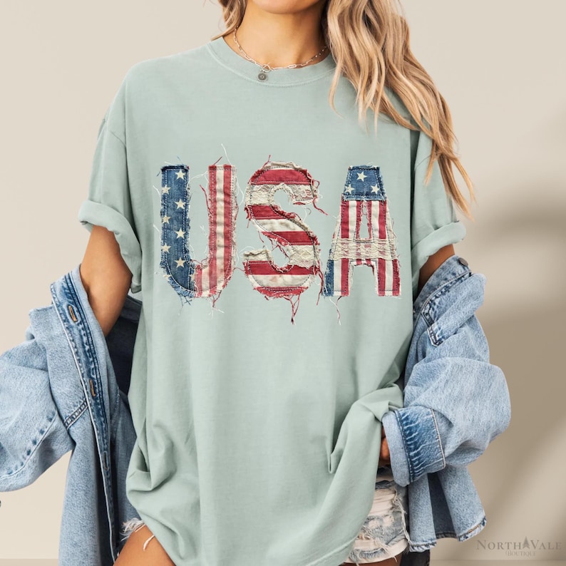 May include: A light green t-shirt with the word "USA" in a distressed, patriotic design. The letters are made to look like the American flag, with red and white stripes and blue stars. The shirt has rolled-up sleeves.