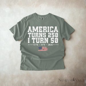 May include: A sage green t-shirt with the text "AMERICA TURNS 250 I TURN 50" in white block letters. Below the text is the American flag and the years 1776, 1976, and 2026. The shirt is a short-sleeved crew neck.