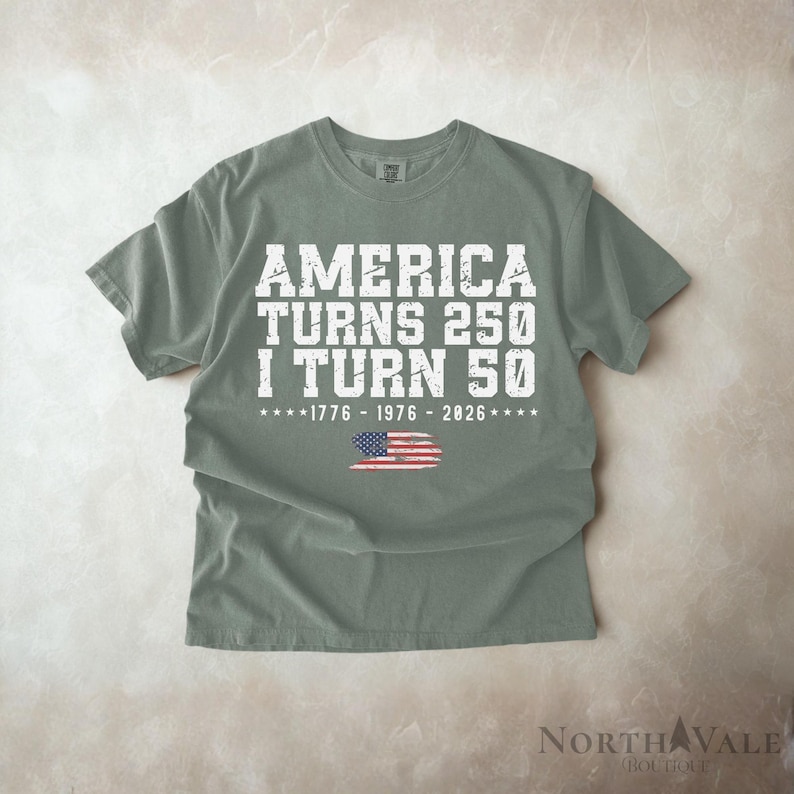 May include: A sage green t-shirt with the text "AMERICA TURNS 250 I TURN 50" in white block letters. Below the text is the American flag and the years 1776, 1976, and 2026. The shirt is a short-sleeved crew neck.