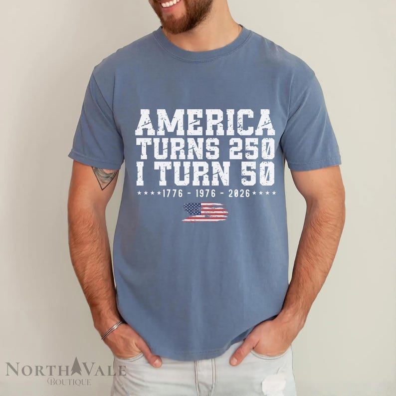 May include: A blue t-shirt with white text that reads "AMERICA TURNS 250 I TURN 50" with the years 1776-1976-2026 and an American flag graphic. The shirt is a crew neck style.