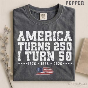 May include: A gray t-shirt with the text "AMERICA TURNS 250 I TURN 50" in white, along with the years 1776, 1976, and 2026. The American flag is at the bottom. The shirt has a Comfort Colors tag.