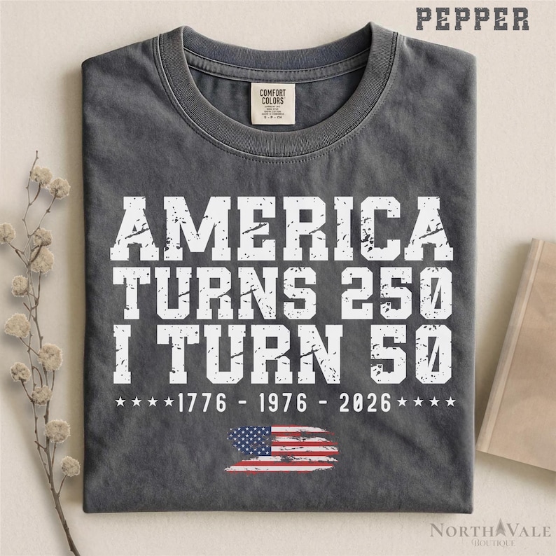May include: A gray t-shirt with the text "AMERICA TURNS 250 I TURN 50" in white, along with the years 1776, 1976, and 2026. The American flag is at the bottom. The shirt has a Comfort Colors tag.