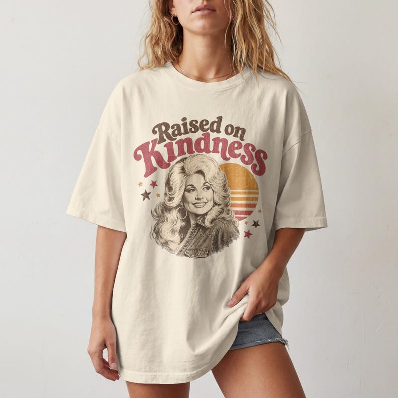 May include: An off-white short-sleeve t-shirt featuring a graphic print of a woman with voluminous hair. The text reads Raised on Kindness in brown and red letters. A circular orange and yellow sunset graphic and small stars surround the portrait.