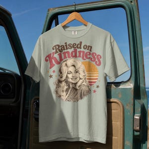 May include: A light sage green short-sleeve t-shirt featuring a graphic print of a woman with voluminous blonde hair. The text "Raised on Kindness" is printed in a curved brown and red font above the portrait, with a sunset and stars.