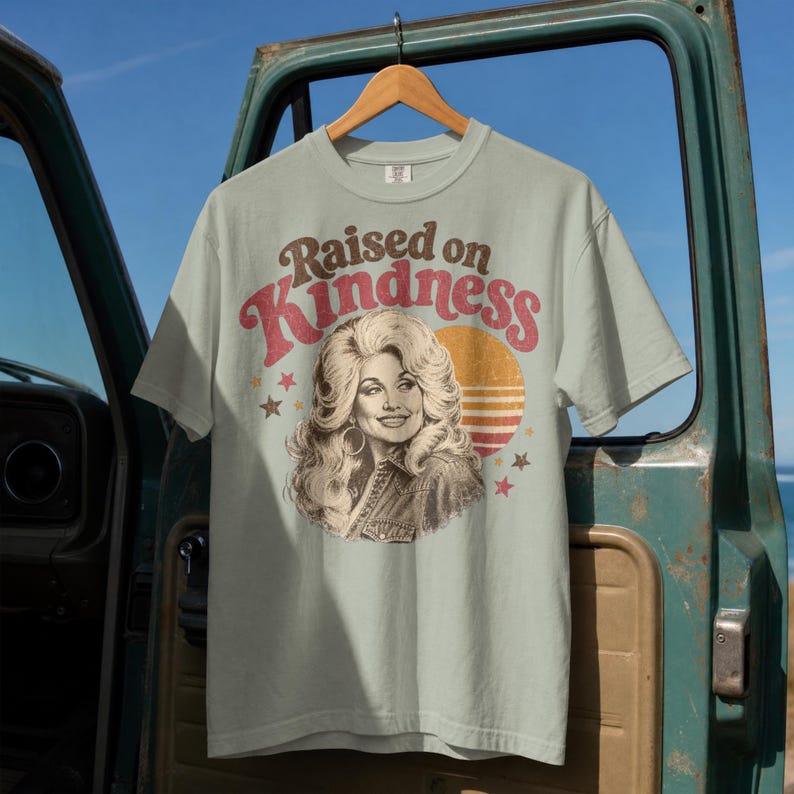 May include: A light sage green short-sleeve t-shirt featuring a graphic print of a woman with voluminous blonde hair. The text "Raised on Kindness" is printed in a curved brown and red font above the portrait, with a sunset and stars.
