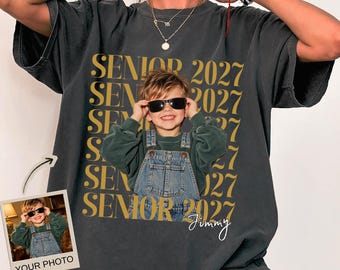 May include: A charcoal gray short-sleeve t-shirt featuring a repeating gold text pattern that reads SENIOR 2027. The center displays a photographic print of a child wearing sunglasses and denim overalls, with the handwritten name Jimmy below.