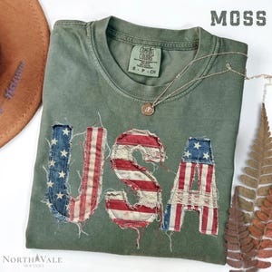 May include: A moss green t-shirt with the word "USA" appliqued on the front in a distressed American flag design. The shirt has a crew neck and short sleeves. A silver necklace with a small pendant is draped across the shirt.